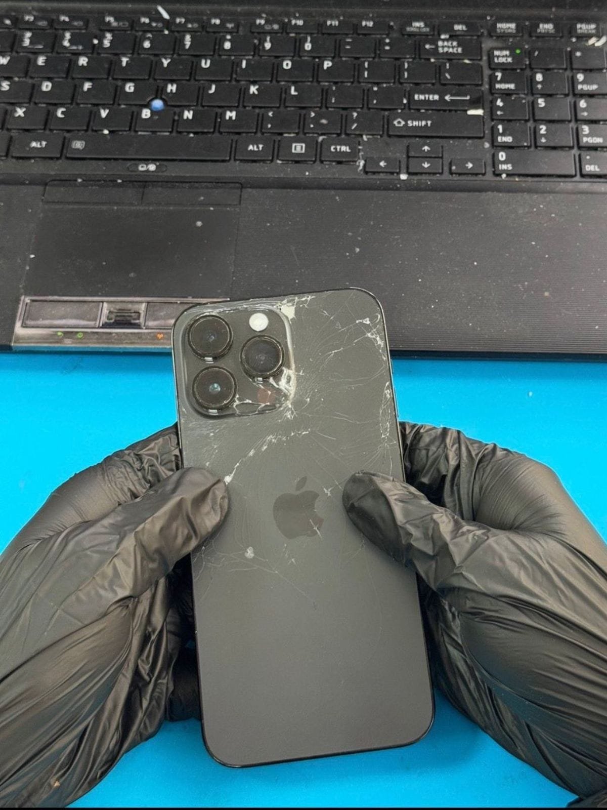 Cracked rear glass on an iPhone 14 Pro Max, impact fracture radiating from beside the camera module, held in gloved hands on a repair bench in Dubai