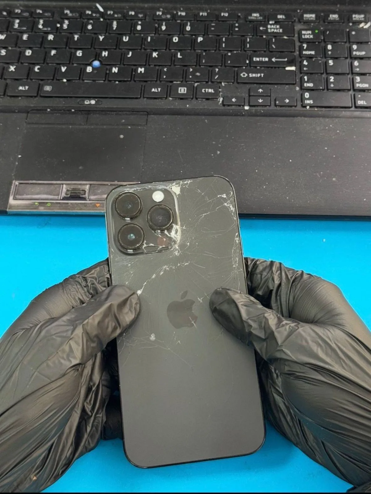 Cracked rear glass on an iPhone 14 Pro Max, impact fracture radiating from beside the camera module, held in gloved hands on a repair bench in Dubai